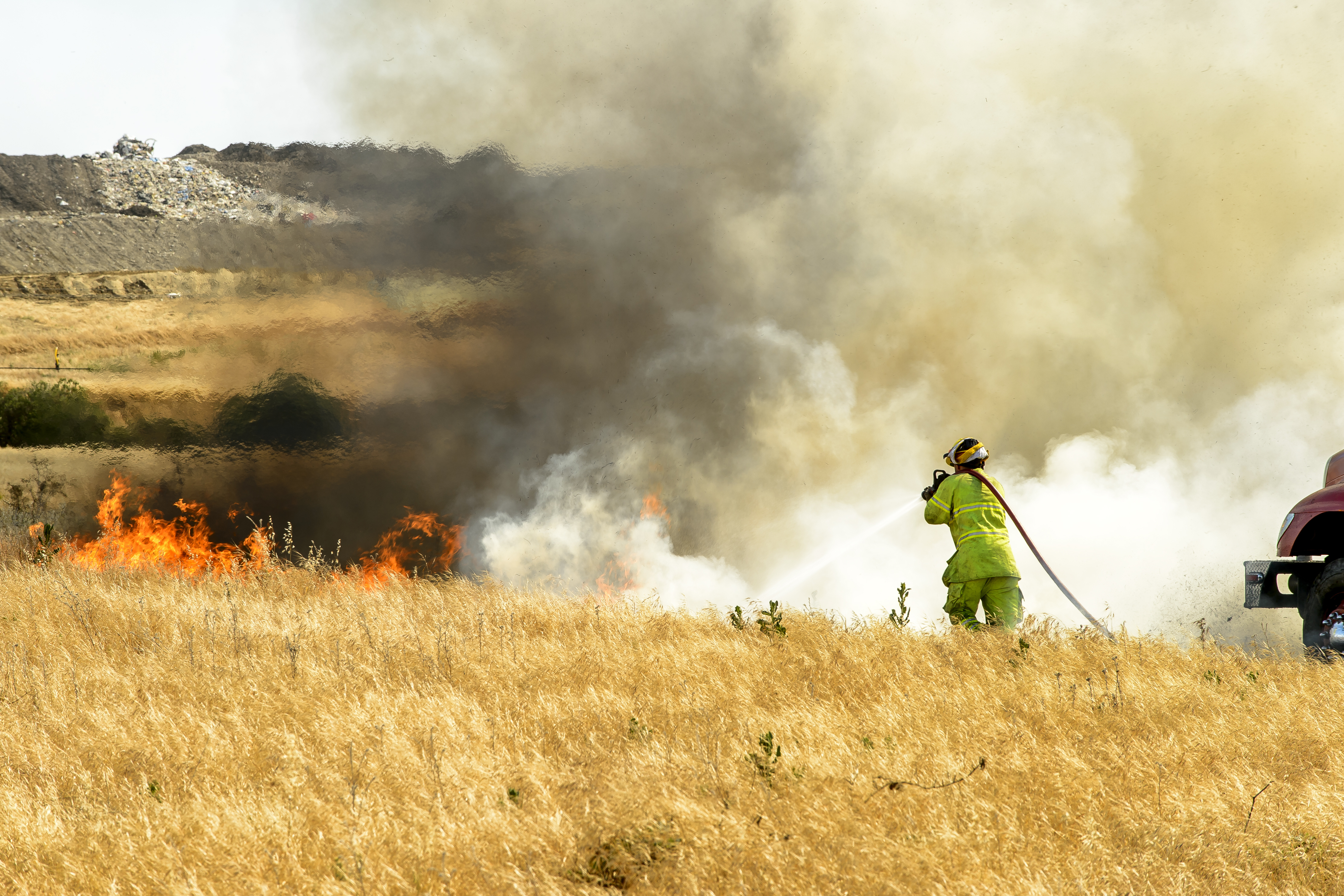 Smoke and flames from dry grass wildfire burning with firefighter hosing down the fire.